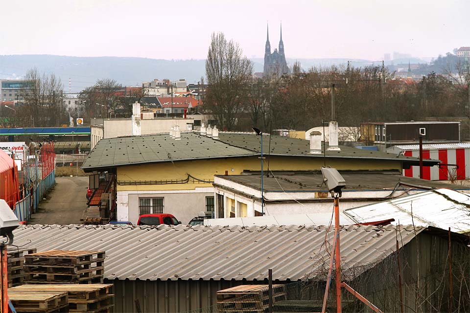 Celkový pohled na areál bývalé stanice Brno-Černovice od východu. Žlutá budova v popředí je nádražní skladiště, ze staniční budovy je vidět jen trojúhelníkový štít mezi stromem a červeným komínem uprostřed snímku.